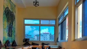 A room with wooden tables and benches, a colorful rug, and a large window with a view of buildings. - Shiva Home Stay (Dharamshala)
