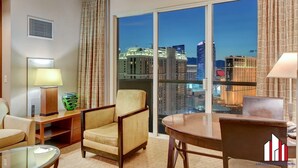 A hotel room with a large window offering a city view, a sofa, a chair, a table, and a lamp. - MGM Signature-30-705 F1 Track & Strip View Balcony (Las Vegas)