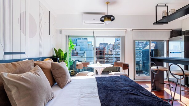 Un dormitorio moderno con cama, una zona de comedor con mesa y sillas, y vistas de la ciudad a través de amplias ventanas.