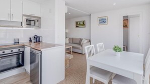 Una cocina compacta con electrodomésticos modernos, un comedor con mesa y sillas blancas, y una sala de estar con sofá y obras de arte en las paredes.