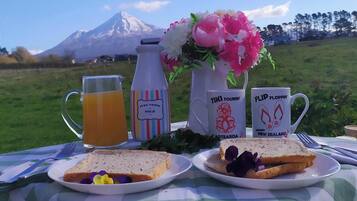 Een picknickopstelling met een kan sap, een melkfles, een vaas met bloemen en twee mokken met tekst, voor een bergpanorama.