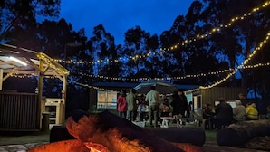 A fire pit with people around it, a covered area, and string lights in the background. - Briggs' Bluff View at Grampians' Edge by Tiny Away (Dadswells Bridge)