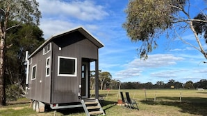 A small, single-story cabin with a front door and windows, situated on a grassy area with trees in the background. - Briggs' Bluff View at Grampians' Edge by Tiny Away (Dadswells Bridge)