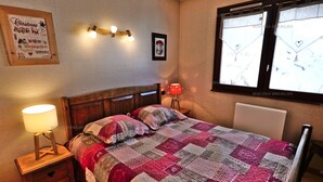 A cozy bedroom with a wooden bed, a quilt, a nightstand with a lamp, a framed Christmas decoration, and a window with a view.
