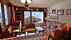 A cozy living room with a wooden interior, a sofa, a coffee table, and a fireplace. There is a large window with a view of a snowy landscape.