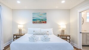 Una habitación de hotel con una cama grande, dos mesitas de noche, un cuadro en la pared y vistas al baño a través de una puerta abierta.