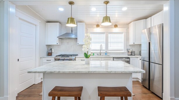 Una cocina moderna con armarios blancos, una isla grande con dos taburetes, electrodomésticos de acero inoxidable e iluminación colgante.