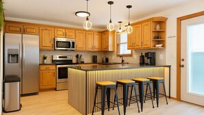 A modern kitchen with wooden cabinets, stainless steel appliances, a central island with stools, pendant lighting, and a view of the outside through a door. - Grand Palace Cottage Escape (Sioux Falls)