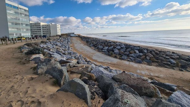 Una passeggiata costiera con una barriera rocciosa, una spiaggia sabbiosa e moderni edifici sullo sfondo.