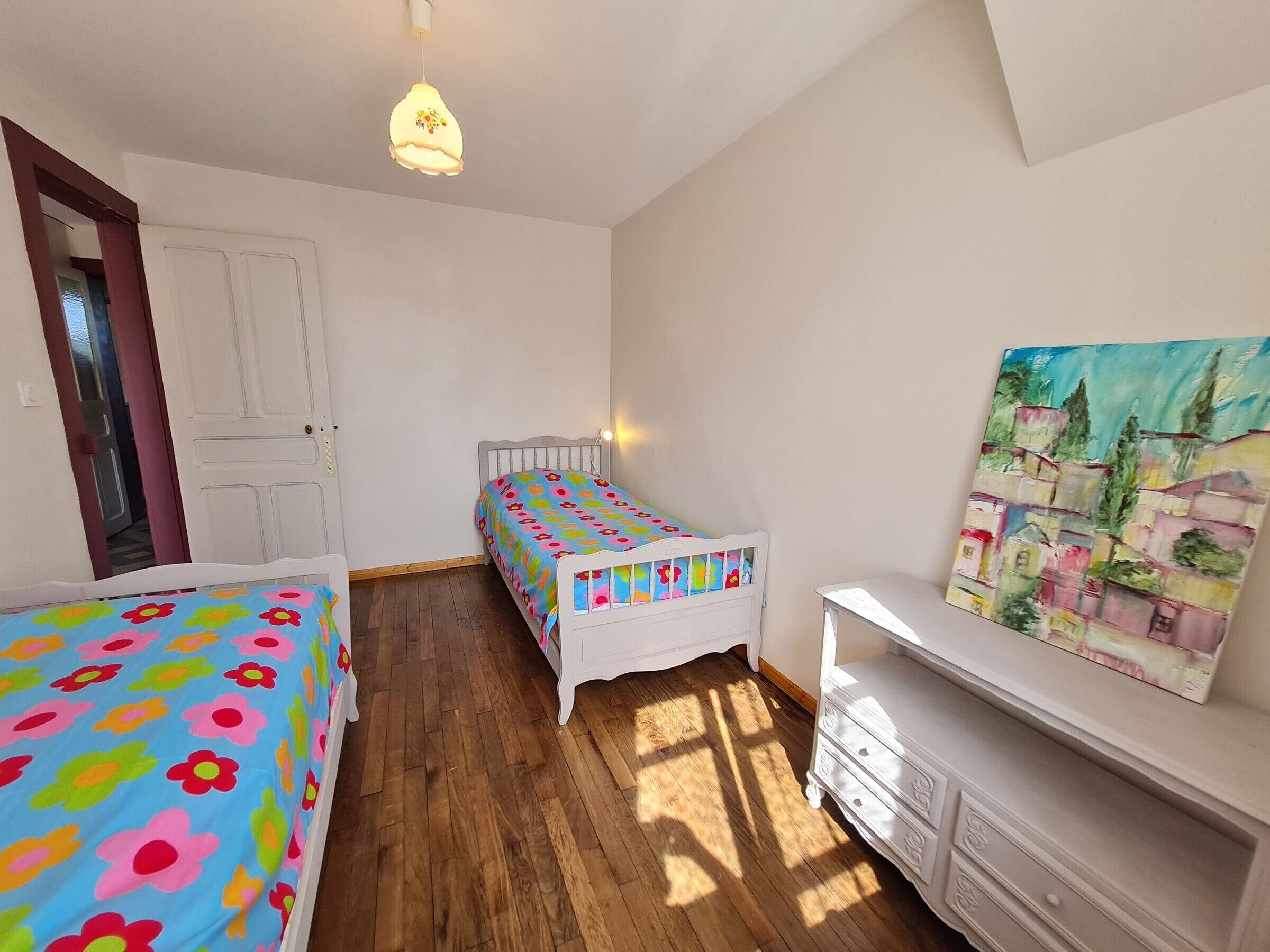 Ein Schlafzimmer mit Holzboden, einem Bett mit bunter Blumenbettdecke, einem Nachttisch, einer Kommode, einem Bild an der Wand und einer Pendelleuchte.