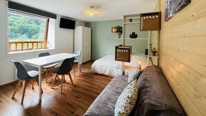 A compact room with a wooden floor, a white desk with chairs, a sofa, a bed, a TV mounted on the wall, and a window with a view of greenery. - Charming studio with parking, mountain view, near Mont-Dore center (Mont-Dore)