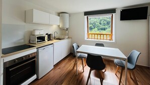A compact kitchen with white cabinets, a microwave, a kettle, and a sink. There's a dining table with two chairs and a window with a view of greenery. - Charming studio with parking, mountain view, near Mont-Dore center (Mont-Dore)