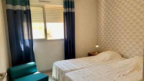 Une chambre avec deux lits, une fenêtre avec des rideaux, une lampe de chevet et un banc de couleur turquoise.