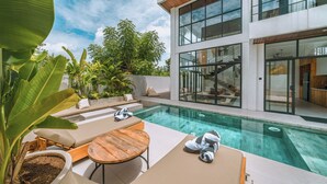 A modern outdoor pool area with lounge chairs, a wooden table, and a staircase leading to an upper level.
