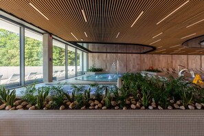 A modern indoor pool area with a wooden ceiling, glass walls, and a hot tub surrounded by plants and rocks.
