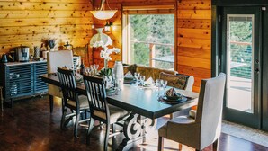 A wooden cabin interior with a dining table set for four, surrounded by chairs, a chandelier, and a window with a view of trees.