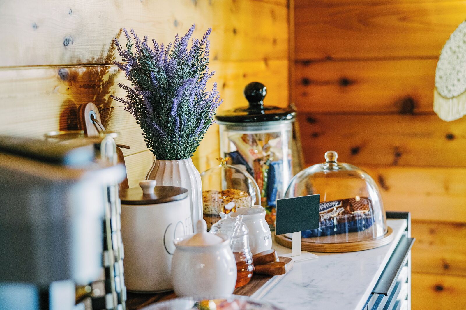 Un plan de travail de cuisine avec une machine à café, un vase de lavande, un pot de confiture et une cloche de verre recouvrant une présentation de chocolats.