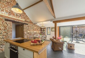 Une cuisine avec un mur en briques, un comptoir en bois et des appareils électroménagers modernes. On y trouve un coin repas avec une table et des chaises, et une vue sur l’extérieur grâce à de grandes fenêtres.
