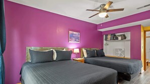 Une chambre avec deux lits, un ventilateur de plafond, un ventilateur fixé au mur et une étagère avec divers objets.