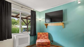 Une pièce aux murs de couleur turquoise, avec une chaise en bois, un téléviseur fixé au mur et une fenêtre avec des rideaux.