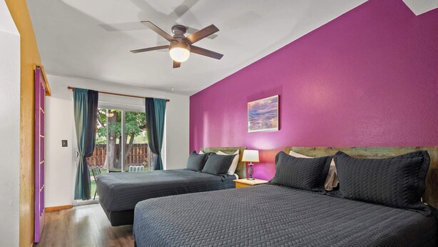 Une chambre avec deux lits, un ventilateur de plafond, une fenêtre avec des rideaux et un mur violet.