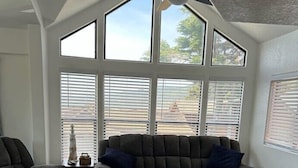 Una sala de estar acogedora con un sofá seccional gris oscuro, una mesita de centro y una alfombra. Hay ventanas amplias con persianas, un ventilador de techo y una zona de cocina con un armario de madera.