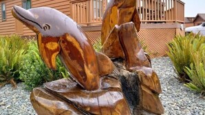 Una escultura de madera con dos cabezas de pez sobre una roca, y una casa de madera al fondo.