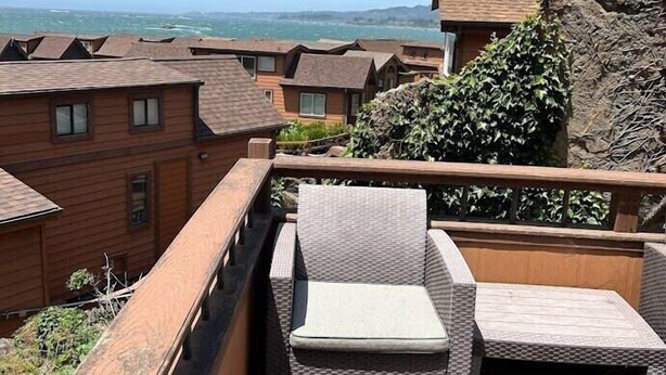 Terraza de madera con mobiliario de mimbre, con vista a una zona costera y varios edificios.