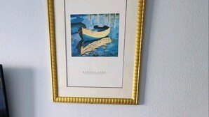 Uma mesa redonda de madeira com quatro cadeiras, um quadro emoldurado de um barco num lago e uma televisão sobre um suporte.