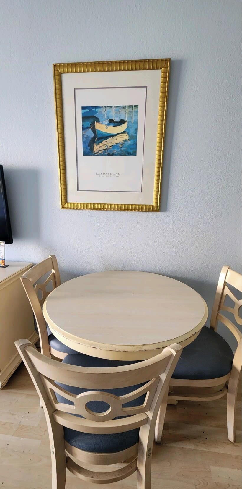 A round wooden table with four chairs, a framed picture of a lake on the wall, and a television on a stand to the left.