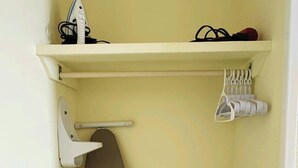 Quarto pequeno com parede bege, teto branco, tábua de passar roupa bege, prateleira branca com secador de cabelo e sapatos, e armário branco pequeno com controle remoto.