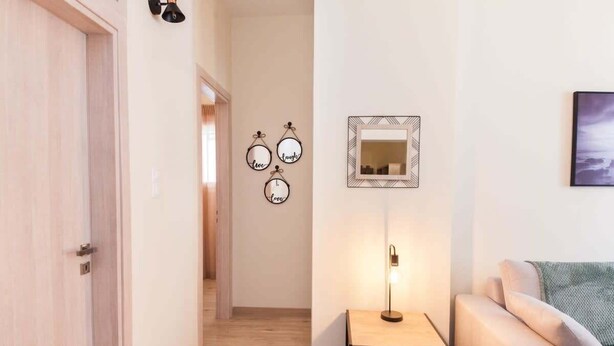 A hallway with a wooden door, a small table with a lamp, a sofa, and wall decorations including framed mirrors and circular signs. - Trendy 2 Bd Apt in Galatsi (Athens)