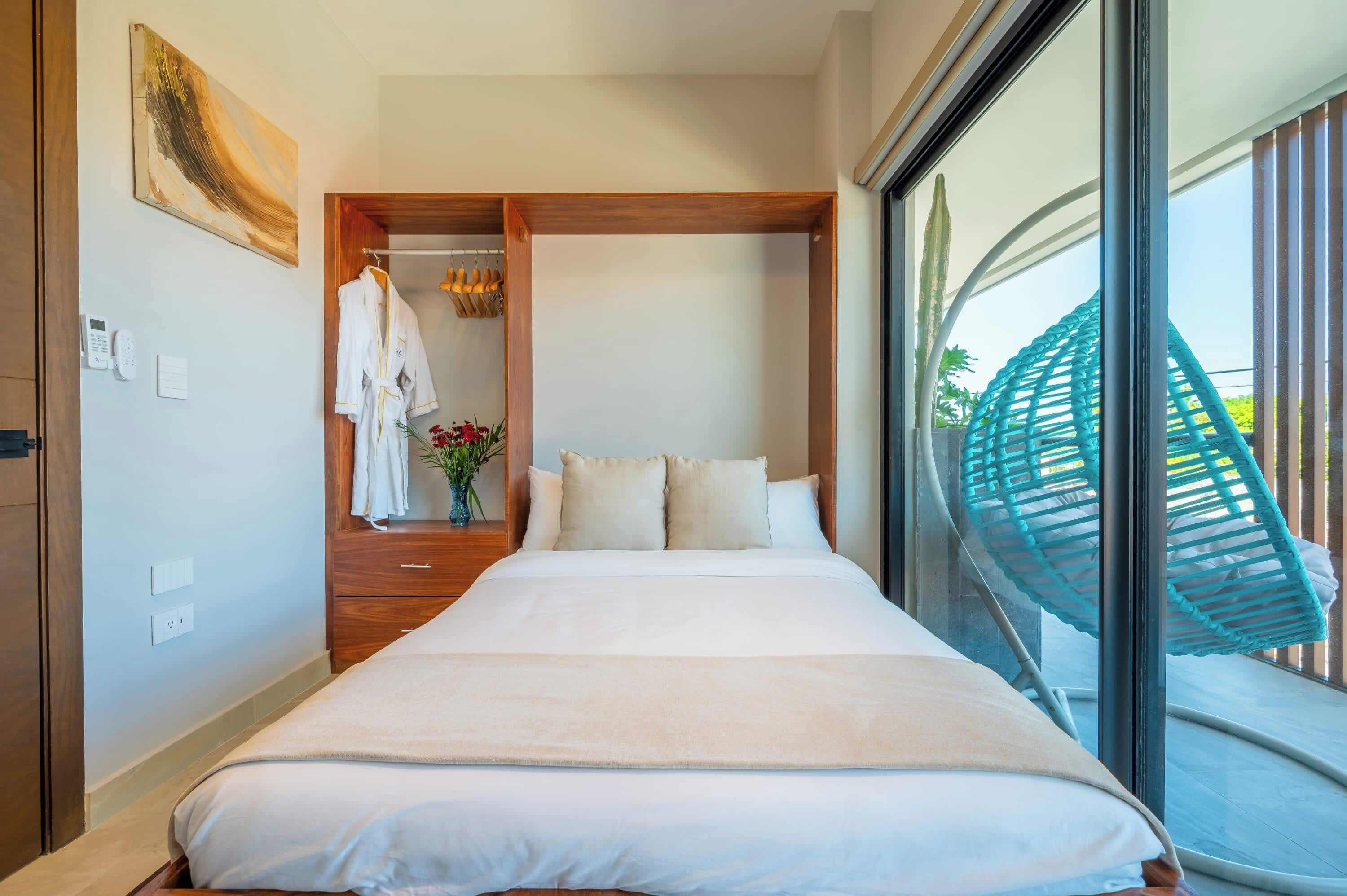 A hotel room with a bed, a wooden wardrobe, a bathroom with a towel rack, and a large window with a view of greenery.