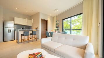 A modern living room with a sofa, a dining area with chairs, a kitchen with a refrigerator, and a large window offering a view of greenery.