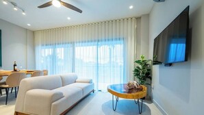 A modern living room with a sofa, a round coffee table, a TV mounted on the wall, and a large window with curtains. - Kippal Holbox 2 Bedroom 2 Baths and a Private Balcony (San Miguel de Cozumel)