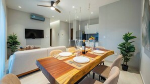 Un comedor moderno con una mesa de madera dispuesta para cuatro personas, un ventilador de techo y un televisor de pantalla plana montado en la pared.