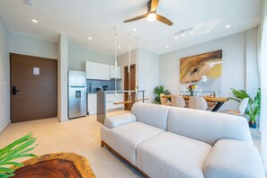 Un salon moderne avec un canapé, un coin repas avec une table et des chaises, une cuisine équipée d’un réfrigérateur et un ventilateur de plafond.