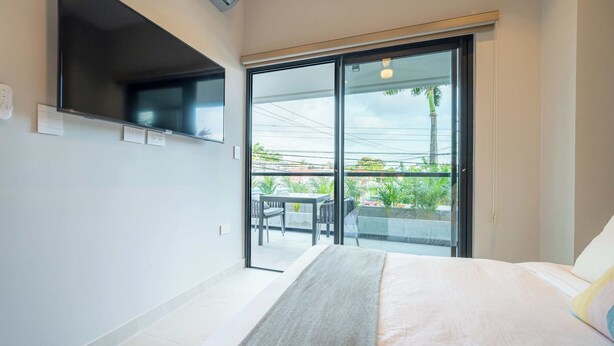 A bedroom with a bed, a TV mounted on the wall, and a sliding glass door leading to a balcony with outdoor furniture.