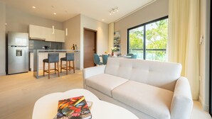 Ein modernes Wohnzimmer mit einer Couch, einem Essbereich mit Stühlen, einer Küche mit Kühlschrank und einem großen Fenster mit Blick auf Grünflächen.