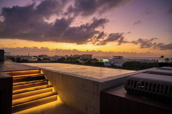 Una terraza en la azotea con vistas a la puesta de sol sobre el agua, una escalera de madera y una zona de barbacoa.