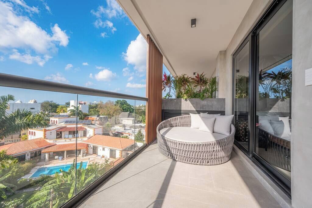 En balkon med et rundt siddeområde, en glasdør og udsigt til en swimmingpool samt bygninger.