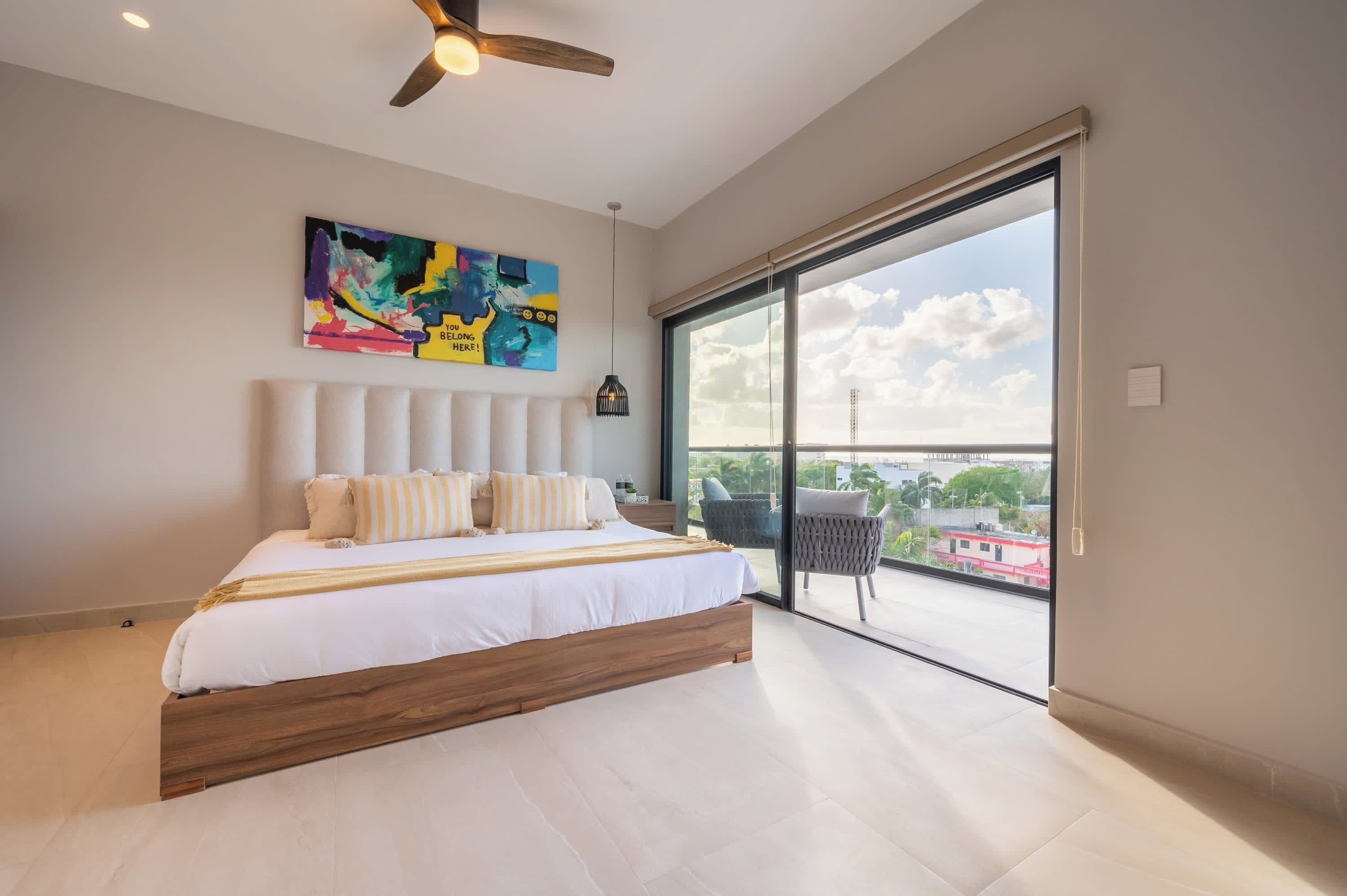 A bedroom with a large bed, a ceiling fan, a colorful abstract painting, and a view of the city through a large window.