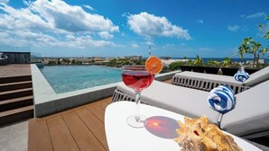 Zona junto a la piscina con vistas claras al océano, un vaso de bebida roja y una concha sobre una mesa blanca.