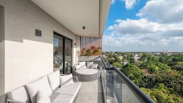A balcony with a sofa and a round seating area, overlooking a cityscape with greenery.