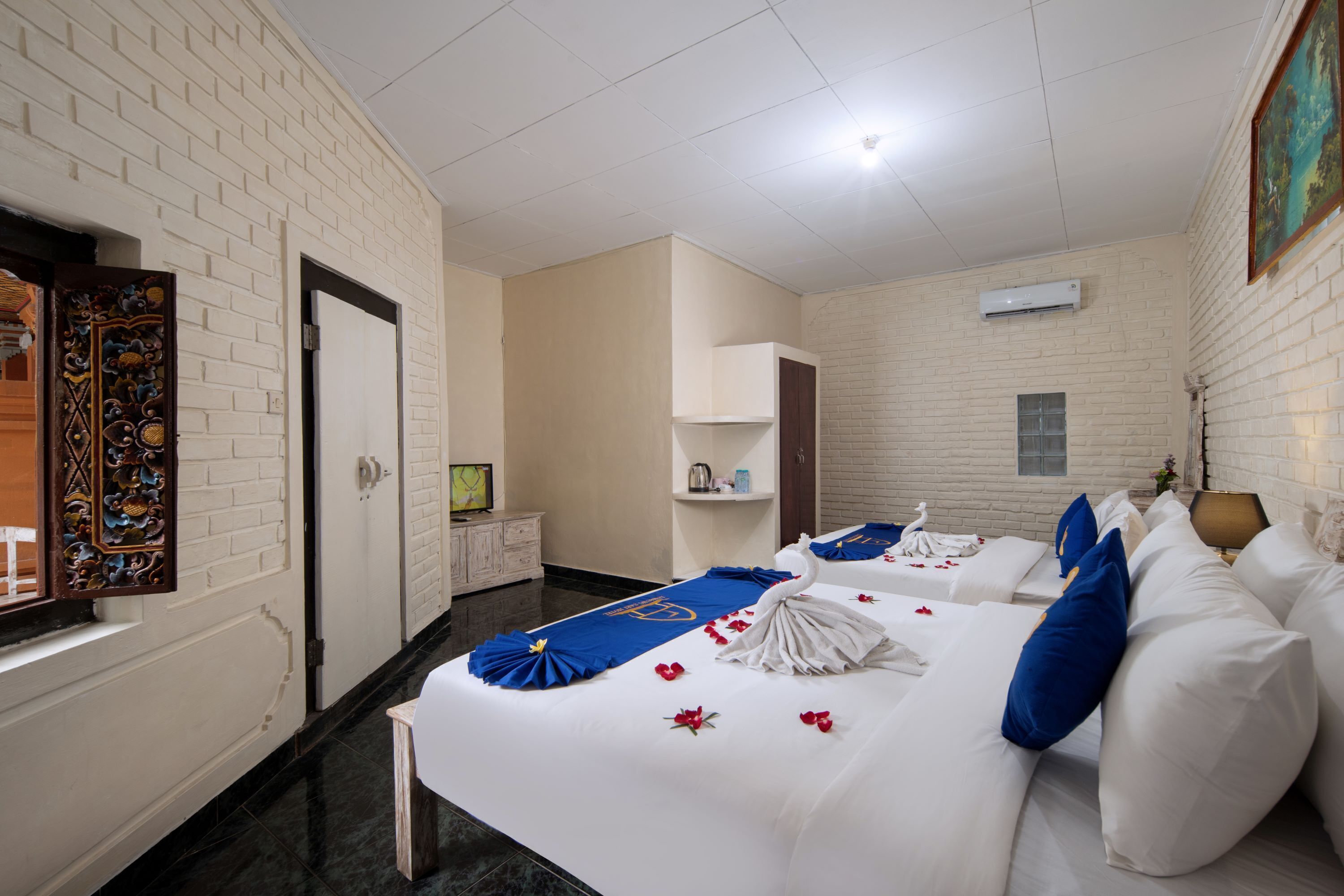 A hotel room with a brick wall, a bed with white bedding and blue pillows, a small bedside table, and a painting on the wall.