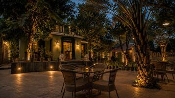 An outdoor reception area with wicker chairs and tables, surrounded by potted plants and trees, under ambient lighting.