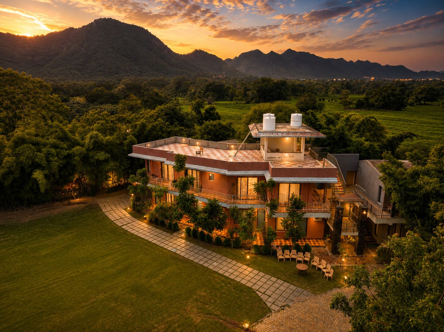 A modern two-story house with a flat roof, surrounded by lush greenery and mountains in the background.