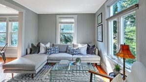 A modern living room with a sectional sofa, a glass coffee table, and large windows offering a view of greenery. - Private Gem in Heart of Downtown Newport  (Newport)