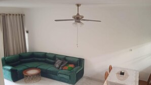 Ein Wohnzimmer mit einem grünen Ecksofa, einem runden Couchtisch, einem Esstisch mit Stühlen und einem Deckenventilator.