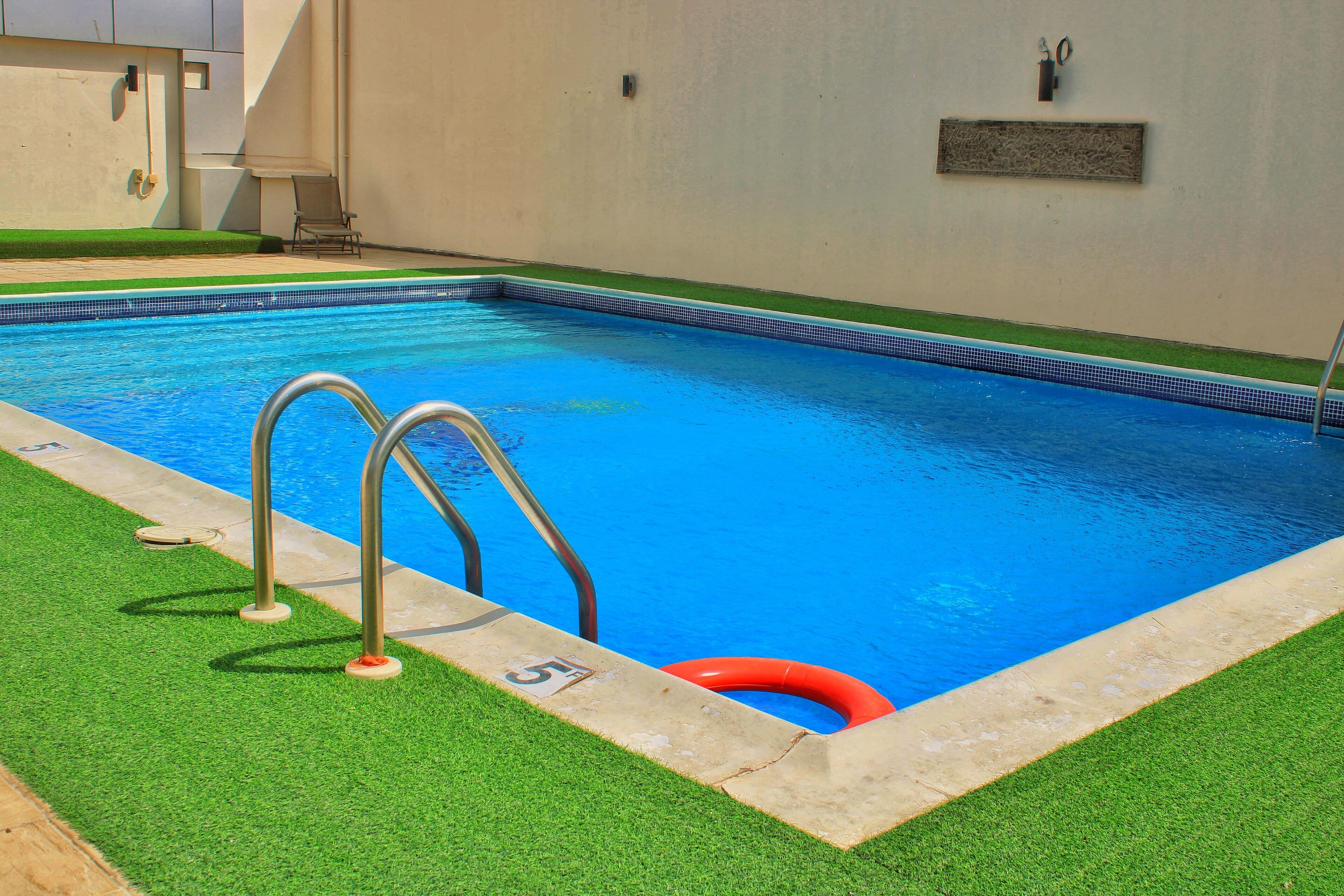 A swimming pool with a clear blue water, surrounded by artificial grass and a concrete edge.